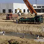 Gru, camion e operai che stanno livellando una gettata di cemento, con un capannone in costruzione sullo sfondo
